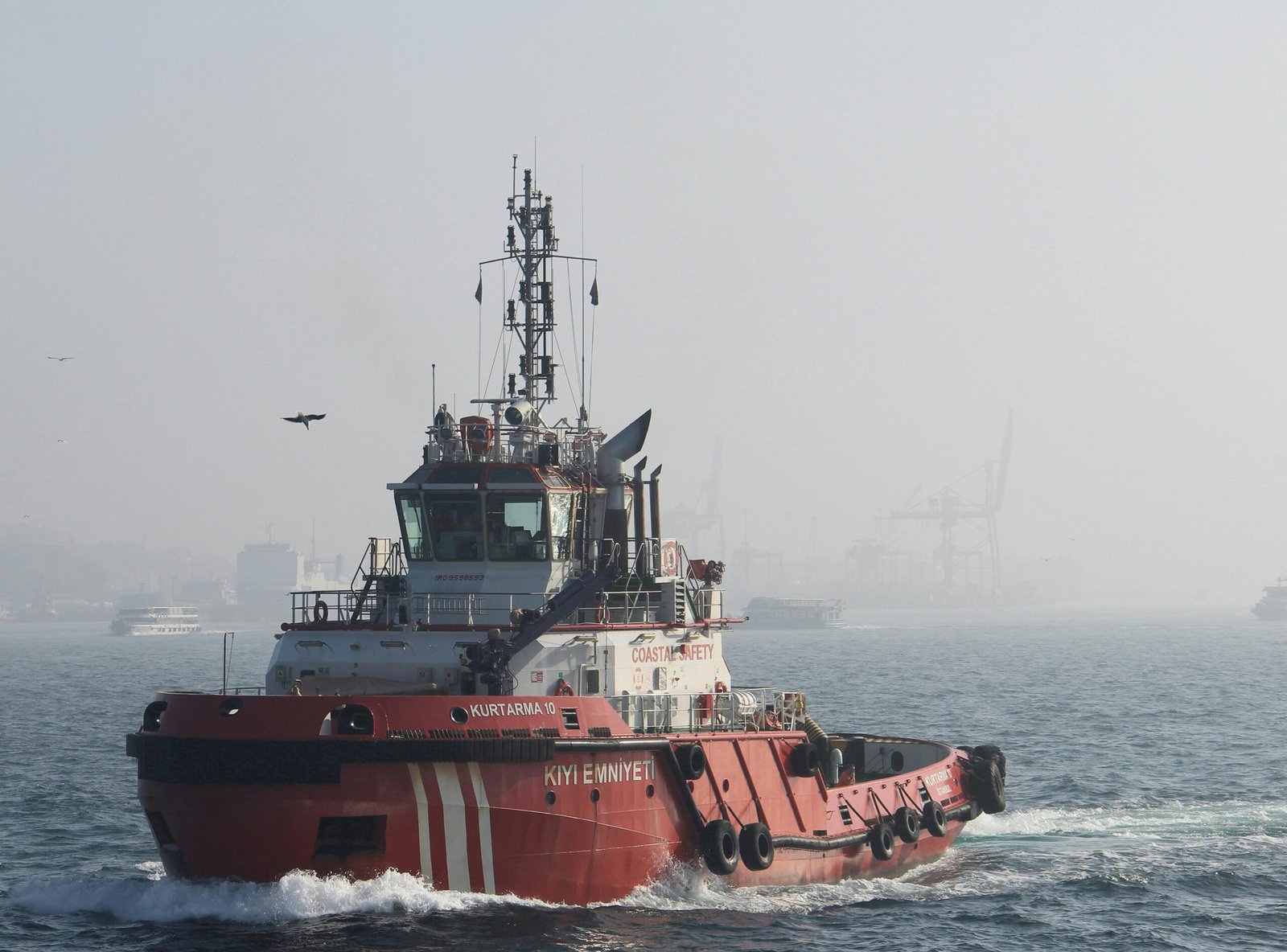 A red tugboat sailing in a misty harbor with cranes in the background.